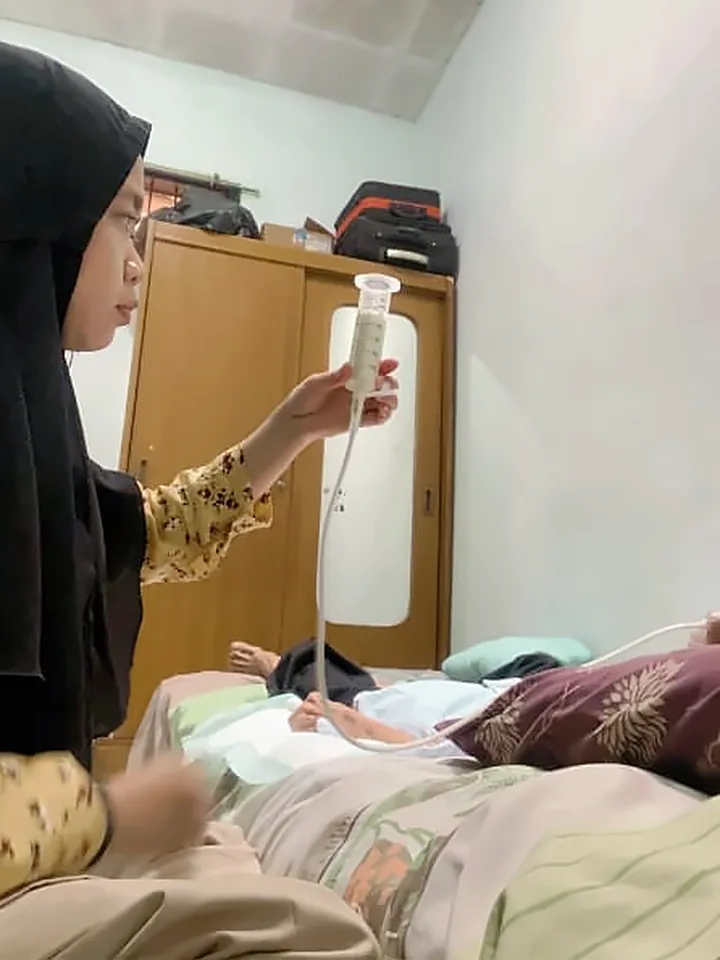 Perawat Orchid Home Care memberi makan pasien lewat selang NGT di rumah, dengan spuit dipegang lebih tinggi dari pasien.